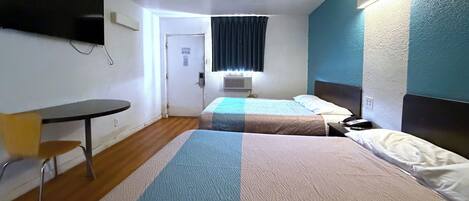 A hotel room with a bed, a chair, a table, a TV mounted on the wall, and a window with curtains.