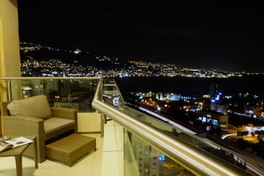 A balcony with wicker furniture and a glass railing overlooking a cityscape at night. - BURJ on BAY Hotel (Kfar Yassine)