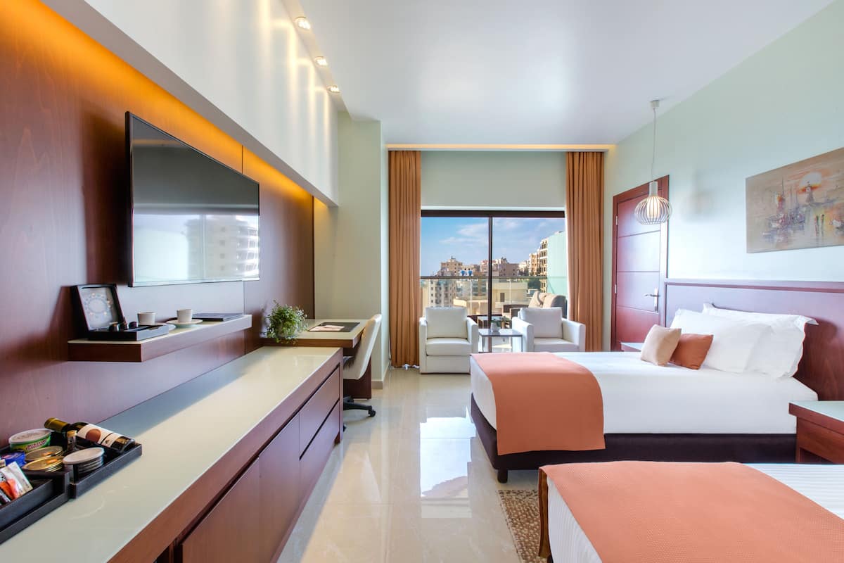 A hotel room with a bed, bedside table, desk, chair, and a large window with a city view.