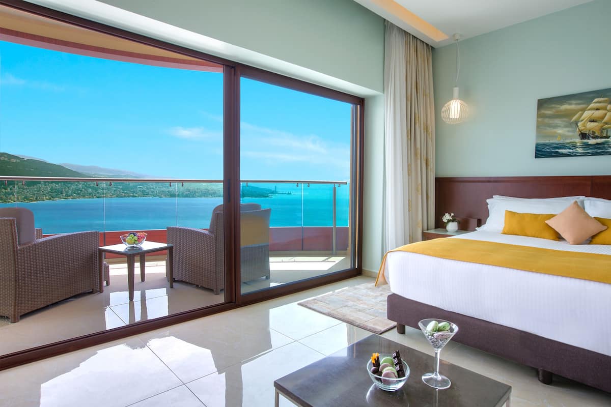 A hotel room with a large bed, a balcony with a view of the sea, and a dining area with a table and chairs.