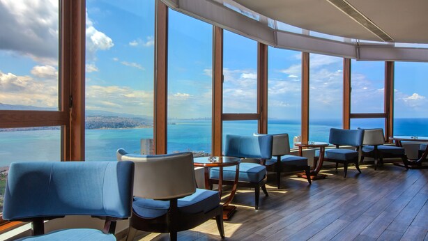 Habitación con amplios ventanales que ofrecen vistas al océano y al paisaje urbano. Hay varias sillas y una pequeña mesa.