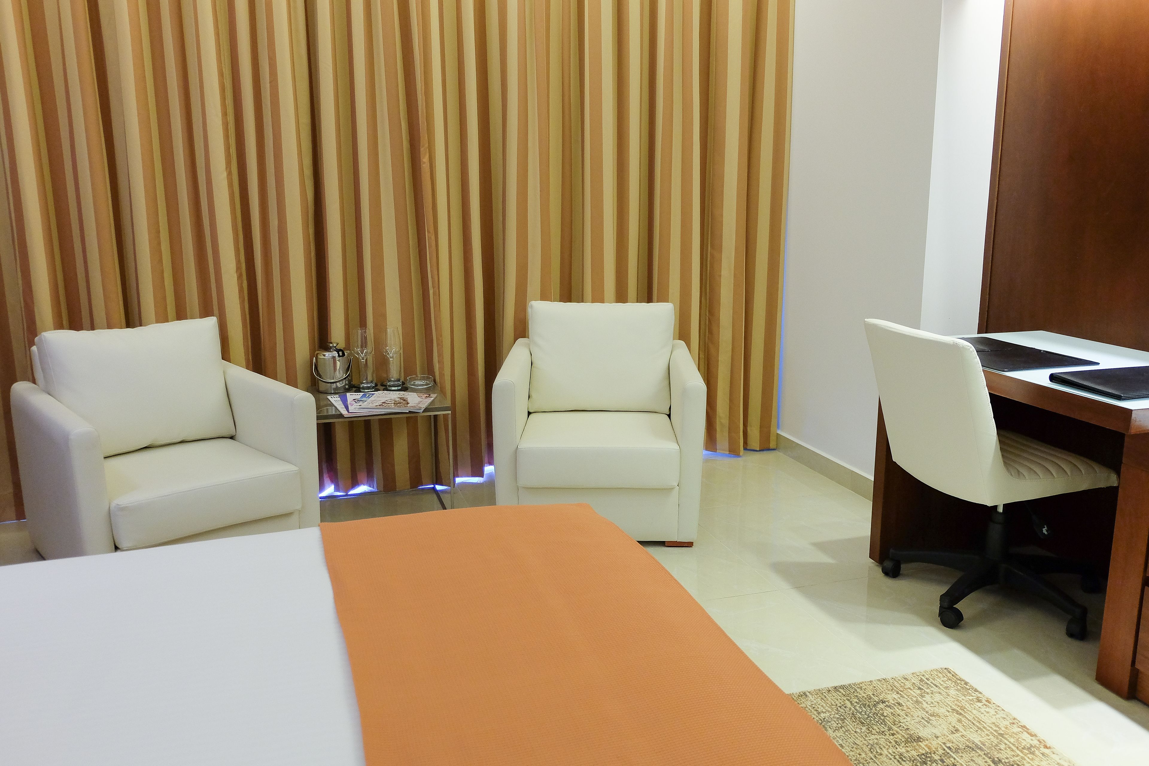 A hotel room with two white armchairs, a wooden desk with a laptop, and a small table with a lamp and glasses.
