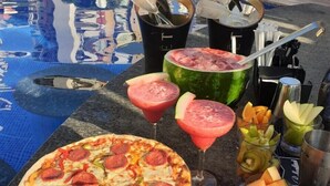 Escena junto a la piscina con pizza, sandía, cócteles y fruta.