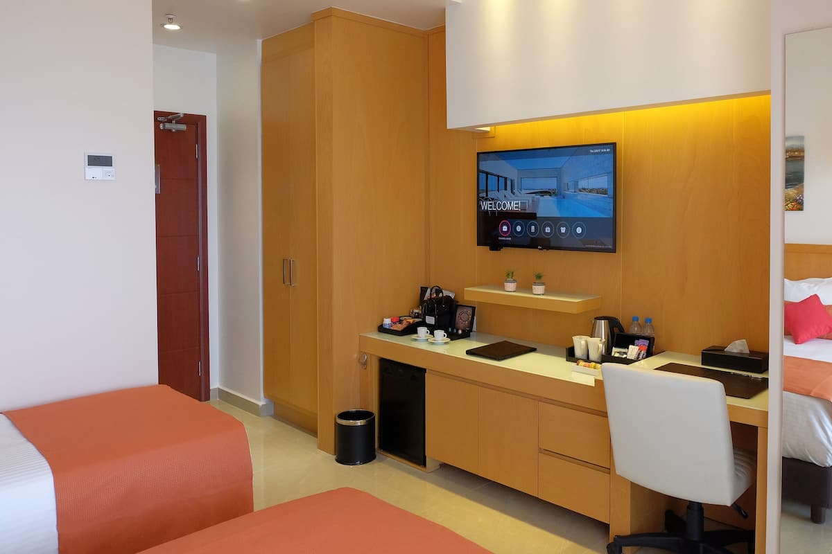 A hotel room with a bed, a desk with a computer, a TV, and a small shelf with toiletries.