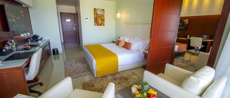 A hotel room with a bed, a desk, a chair, a small table with fruit, and a painting on the wall.