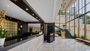 Zona de recepción de un hotel con mostrador de mármol, una escalera y amplias ventanas con vistas al exterior.
