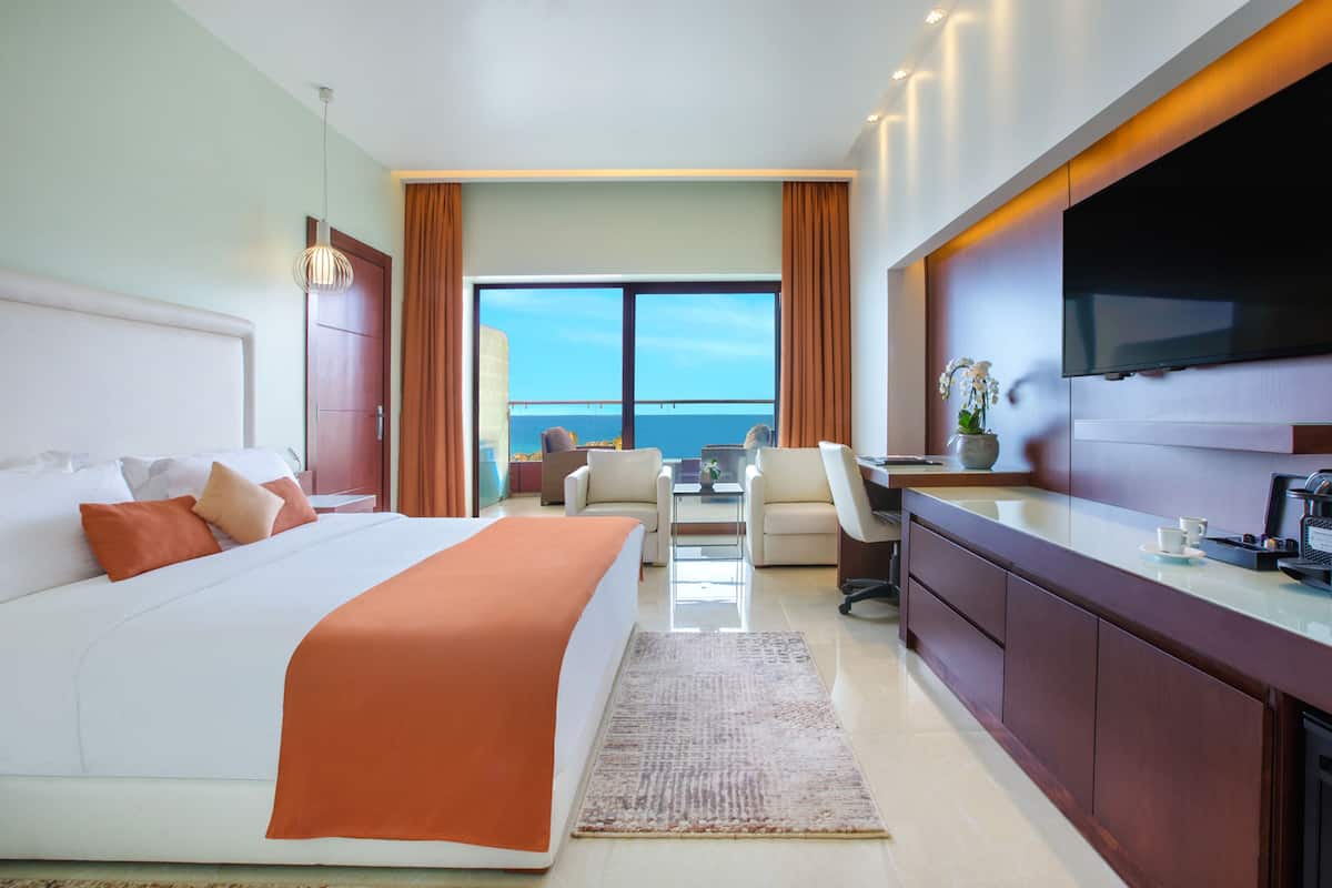 A hotel room with a large bed, a desk, a TV, and a view of the ocean.