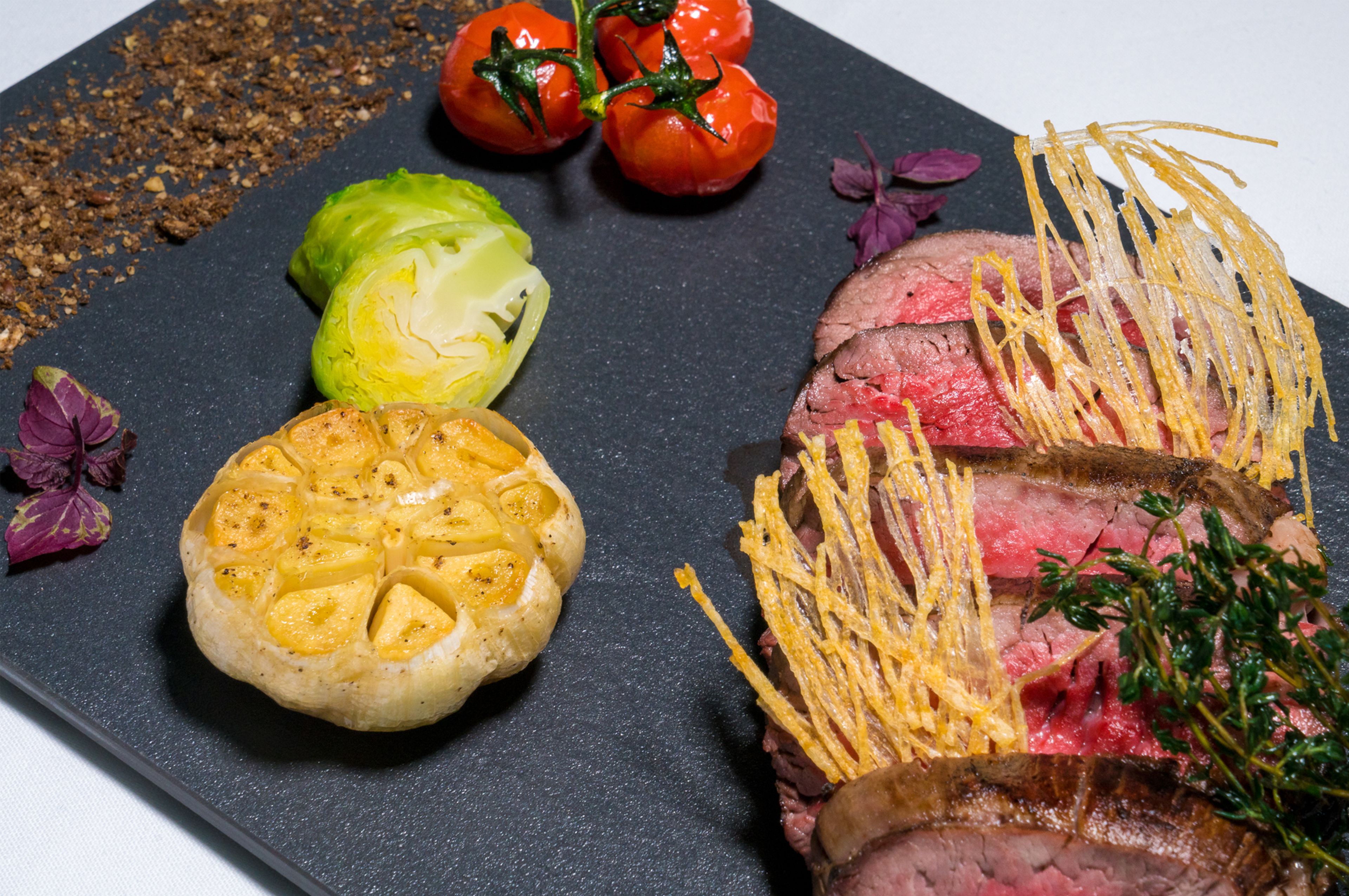 Una tabla de pizarra negra con una porción de carne cubierta de queso rallado, un plato horneado redondo con costra dorada y dos tomates en su vid.