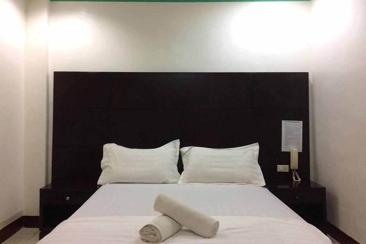 Una habitación de hotel con una cama grande, dos almohadas blancas, un cabecero oscuro y una mesita de noche con una lámpara.