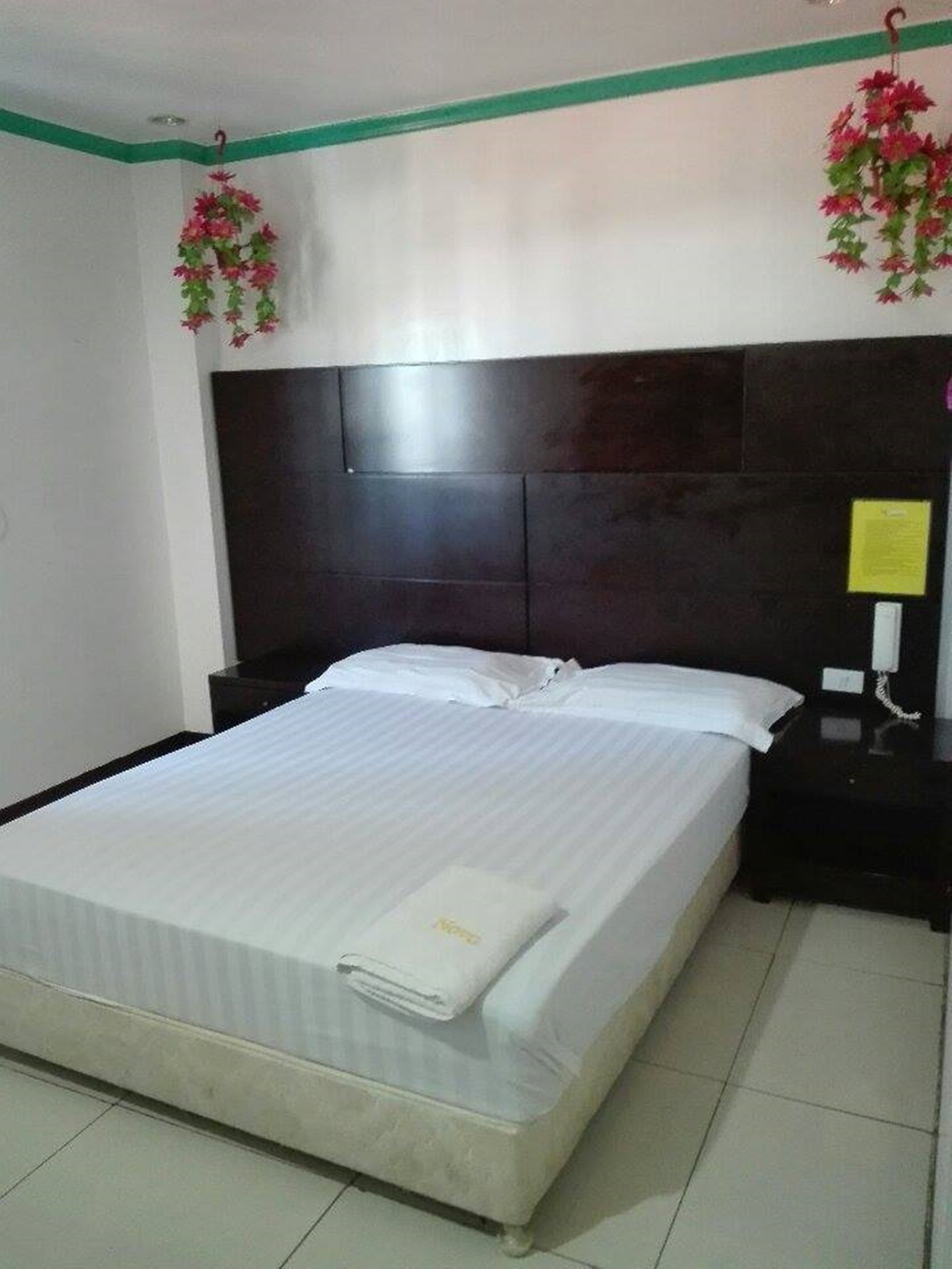A neatly made bed with white linens and a folded towel, a dark headboard, a nightstand with a phone, and two hanging flower arrangements.