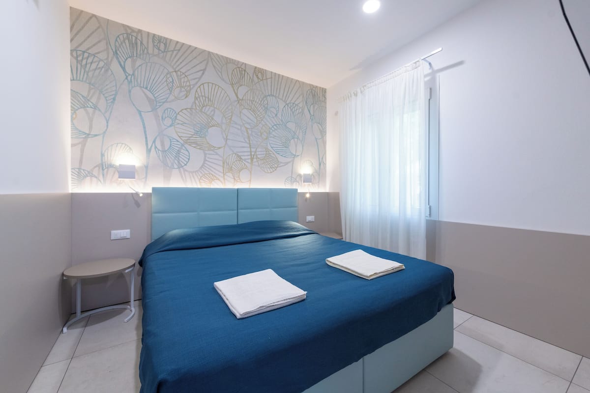 Une chambre moderne avec une couvre-lit bleue, deux serviettes blanches soigneusement pliées, une petite table de chevet et un mur orné d’un motif décoratif.