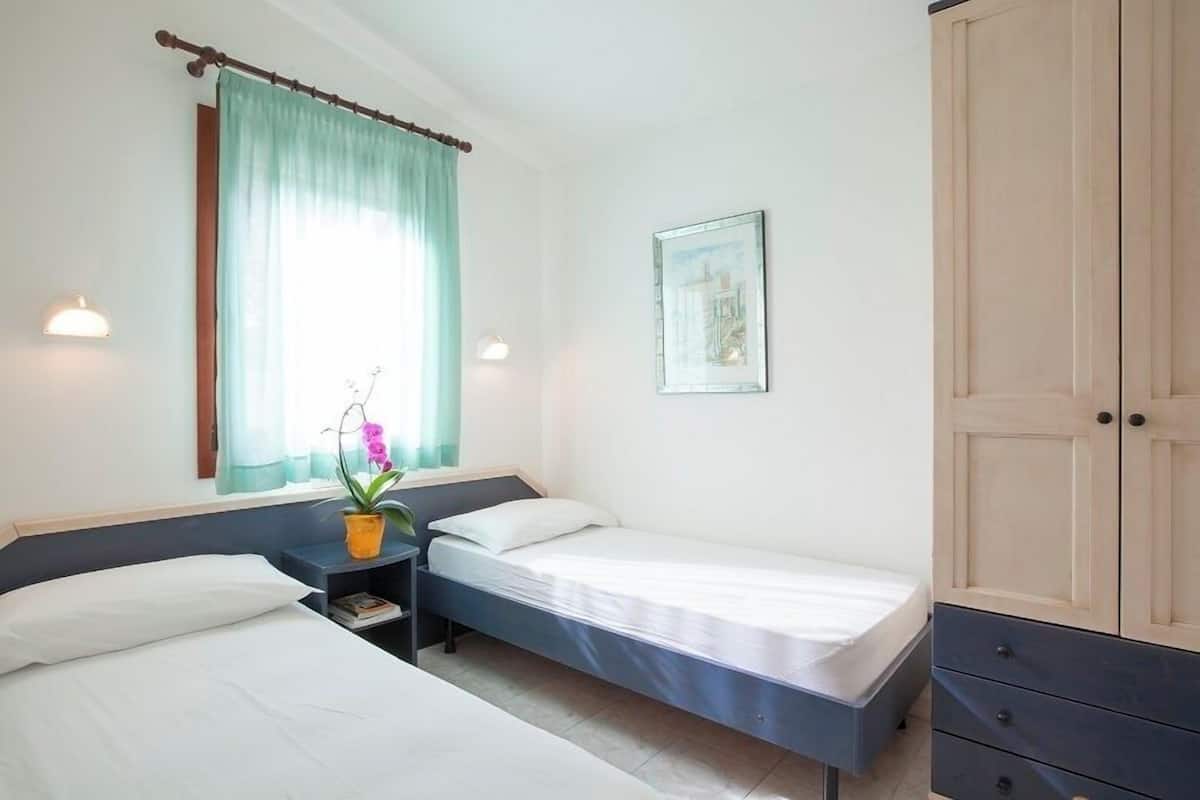 Une chambre avec deux lits simples, une table de chevet avec une lampe, une fenêtre avec des rideaux, un tableau encadré au mur et une armoire.