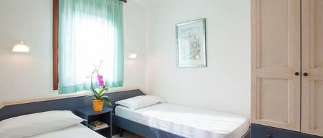 Un dormitorio con dos camas individuales, una mesita de noche con lámpara, una ventana con cortinas, un cuadro en la pared y un armario.