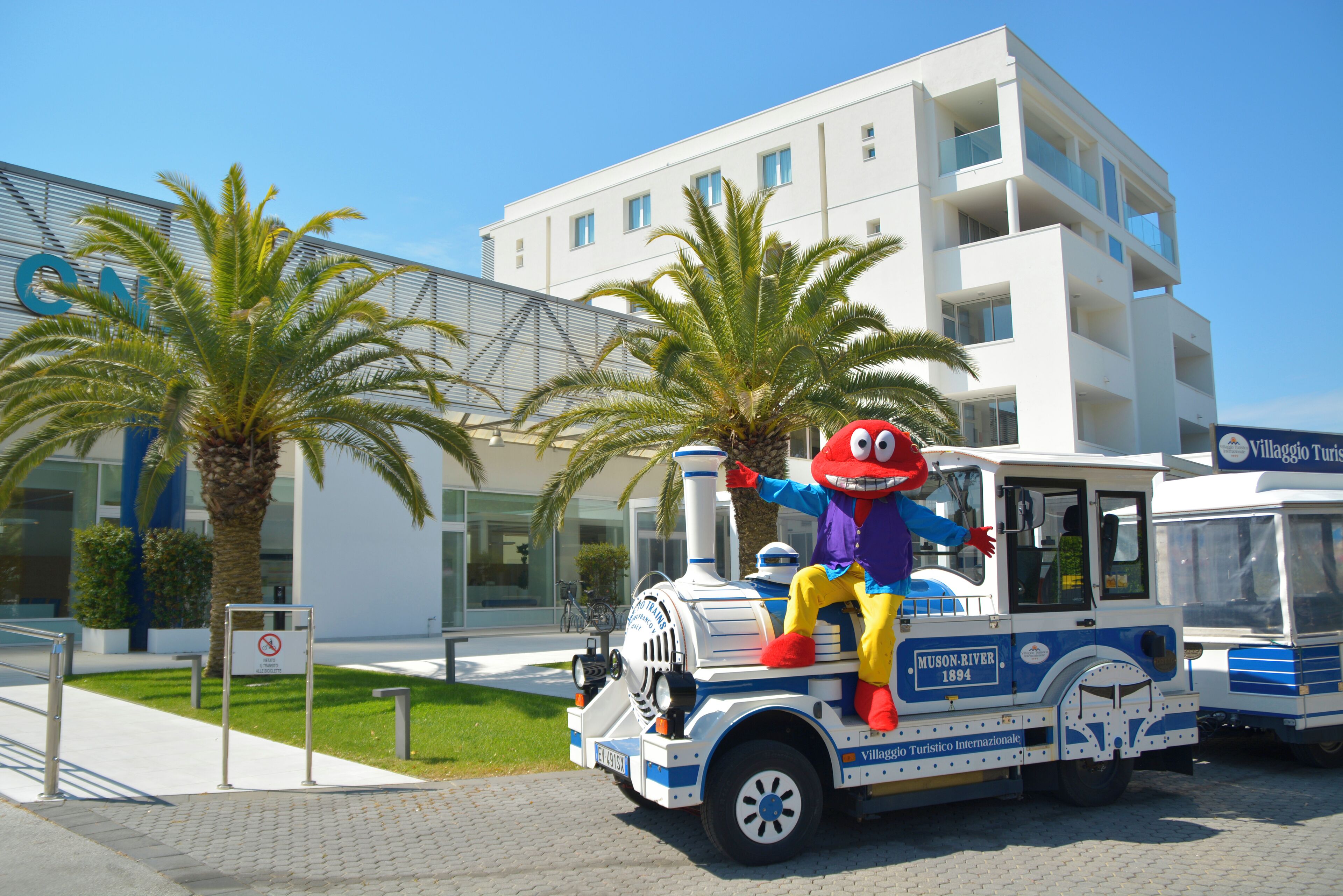 Une mascotte se trouve sur un véhicule décoré selon un thème particulier, devant un bâtiment sur lequel est apposée une enseigne indiquant « Villaggio Turistico Internazionale ».