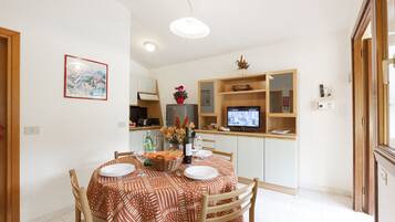 Un comedor con una mesa lista para cuatro personas, una cocina con televisor y un mueble de madera con un jarrón de flores.