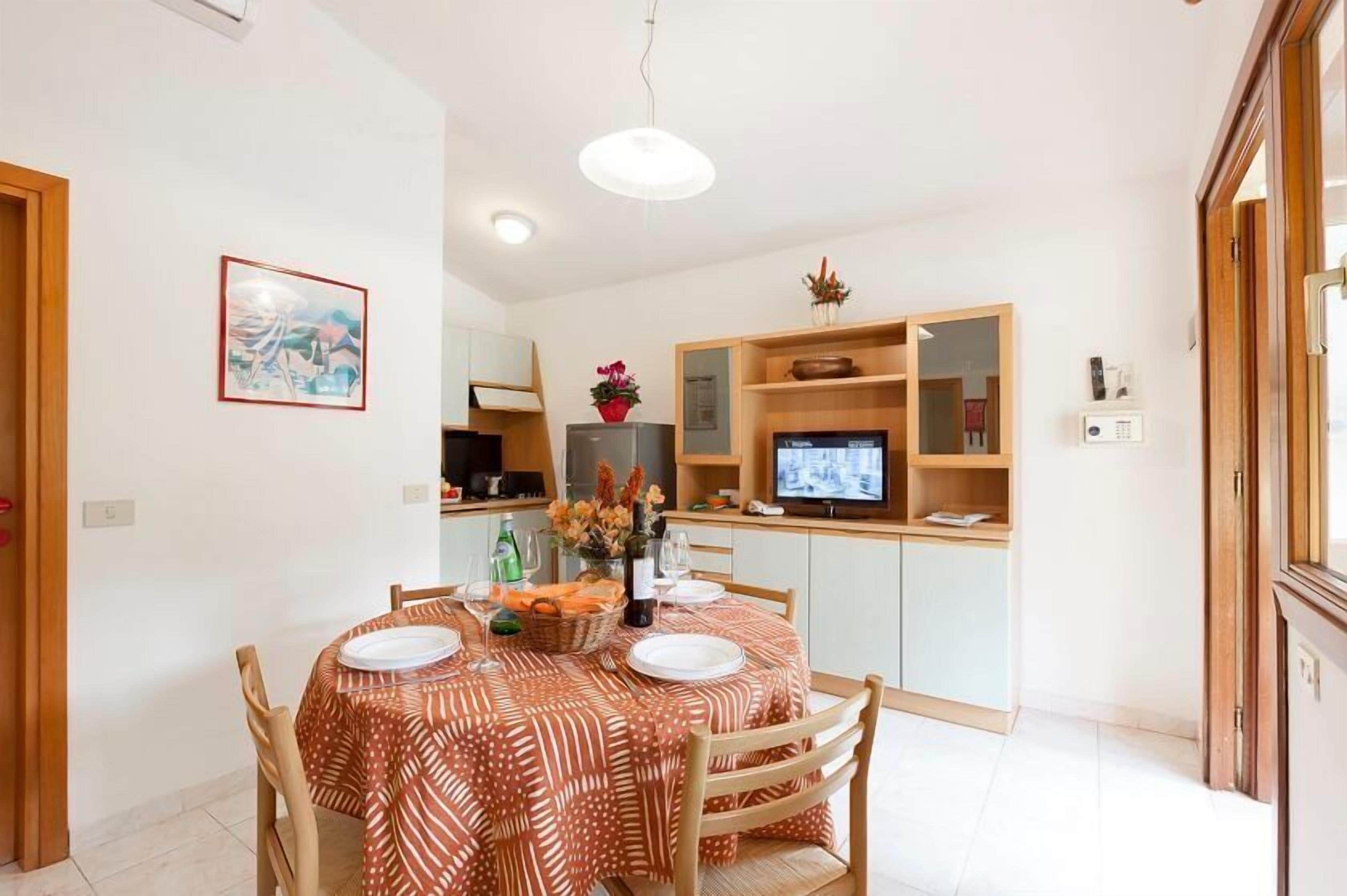 A dining area with a round table set for four, a television on a stand, and a kitchen area with cabinets and a refrigerator.