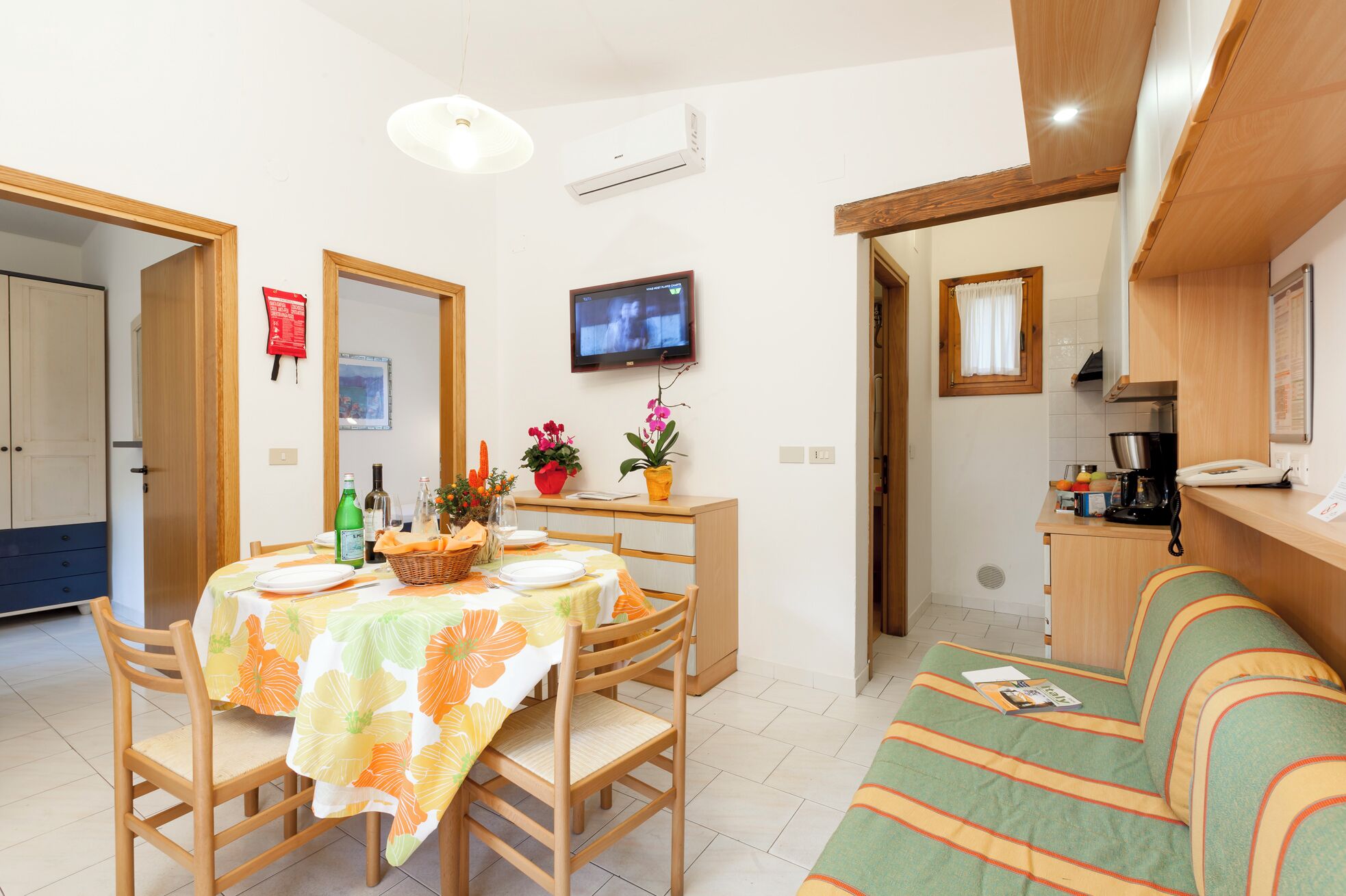 A dining area with a table set for four, a television mounted on the wall, and a kitchenette in the background.
