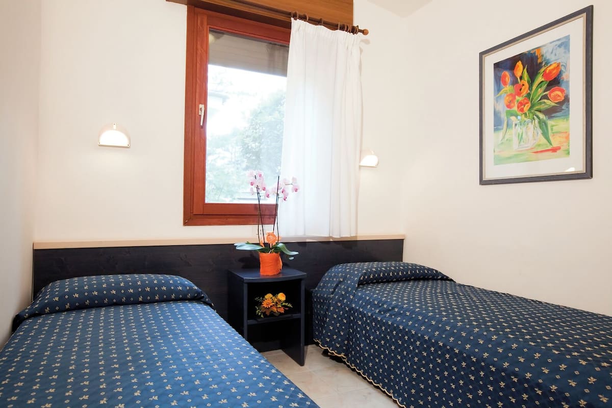 Une chambre avec deux lits, une fenêtre avec des rideaux, une petite table sur laquelle est posé un vase de fleurs, et un tableau accroché au mur.