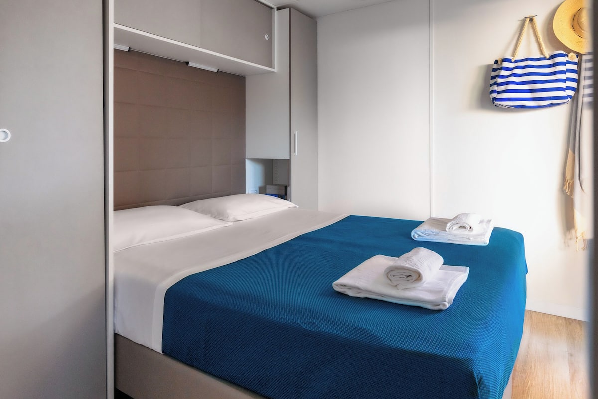 Un lit bien fait, avec une couvre-lit bleue et des oreillers blancs, une petite table de chevet avec une lampe, et un sac à rayures accroché au mur.