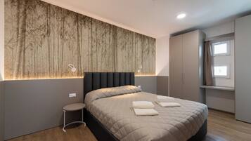 A modern bedroom with a large bed, a small bedside table, a wall-mounted light, and a window with curtains.