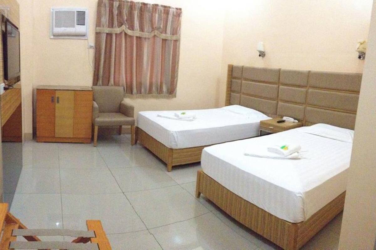 A hotel room with two single beds, a wooden wardrobe, a desk, a chair, and a window with curtains.