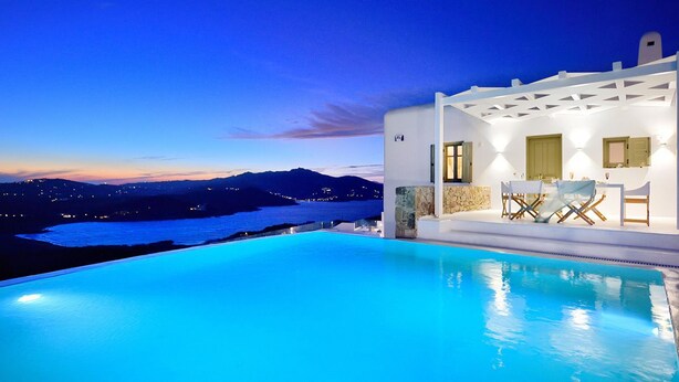 A poolside area with a view of the sea and mountains at dusk.