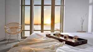 A bedroom with a large bed, a wicker chair, a tray with glasses and a vase, and a view of the sunset through the windows.