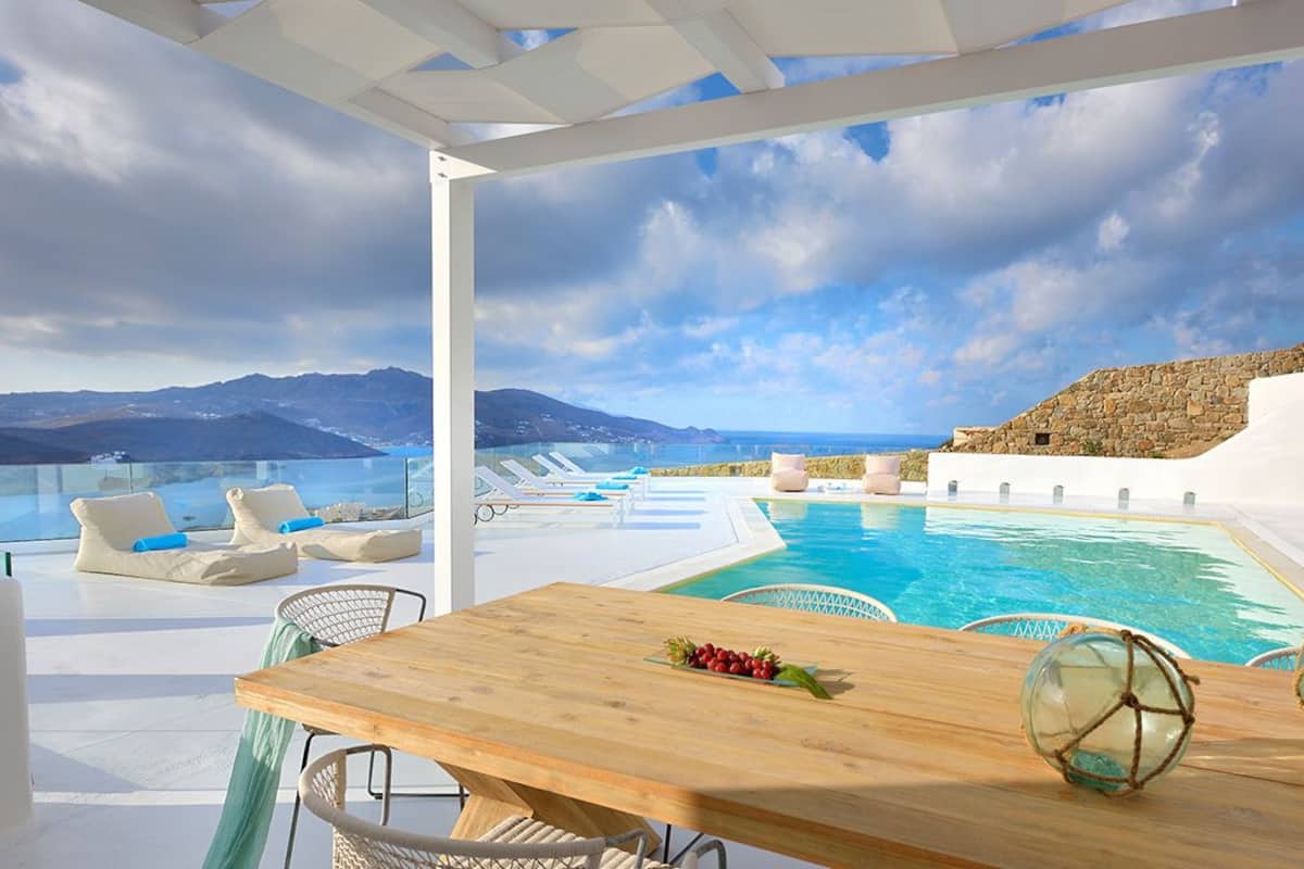 A poolside area with a wooden table, lounge chairs, and a view of the sea and mountains.