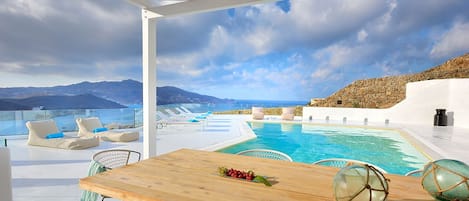 A poolside area with a wooden table, lounge chairs, and a view of the sea and mountains.