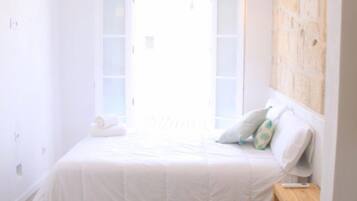 A bedroom with a large bed, a wooden nightstand, a wall-mounted lamp, and a window with a view.