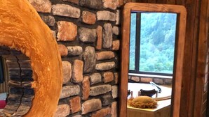 Un interior rústico con una pared de piedra, una mesa redonda de madera con una taza y un espejo que refleja una habitación con una ventana.