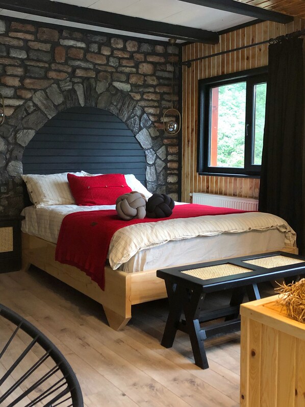 A bedroom with a stone wall, a wooden bed, a window with a view of greenery, and a small wooden table. - Zilkale Butik Otel (Camlihemsin)