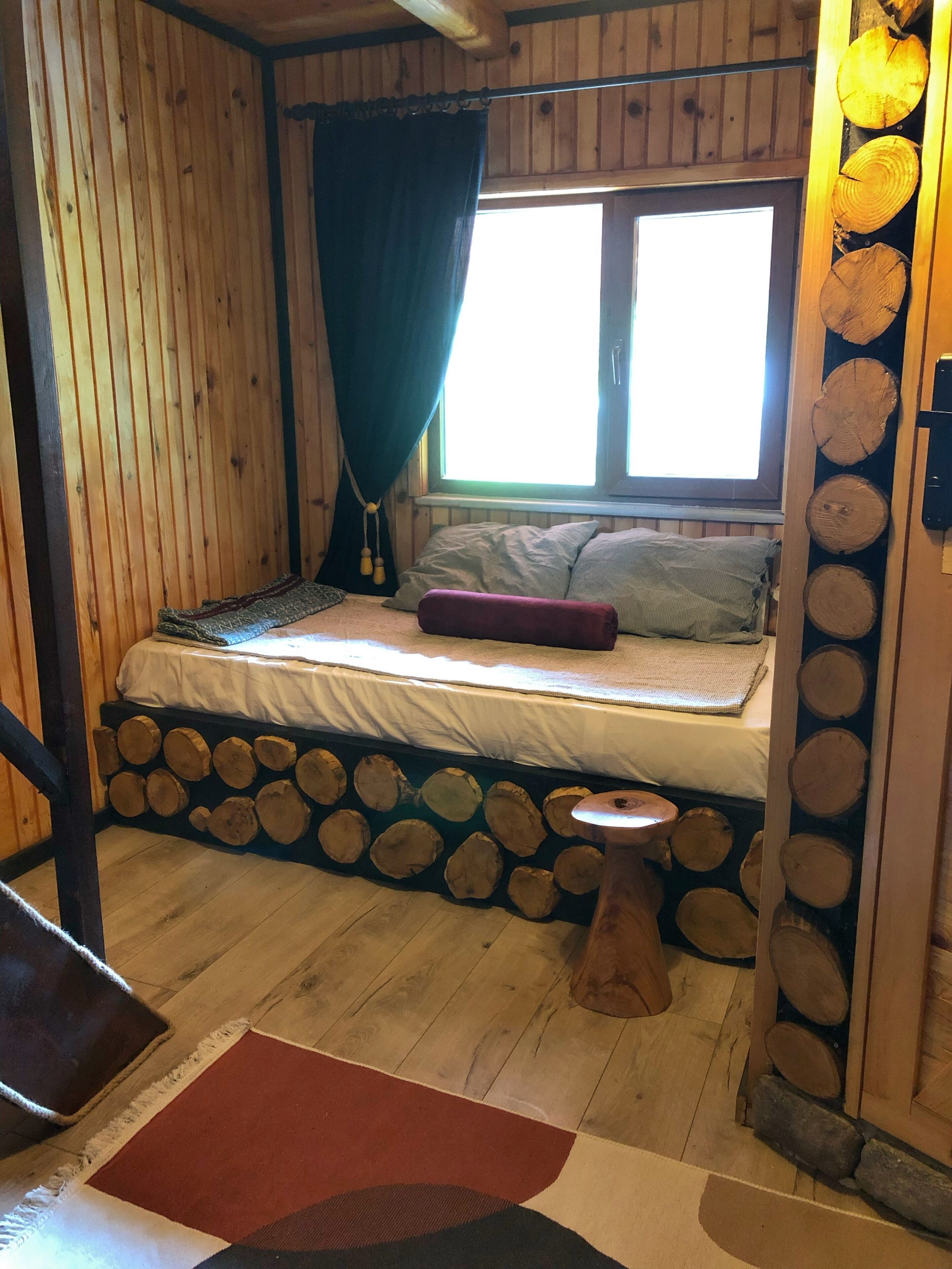 A cozy cabin room with a bed, a window with curtains, a small stool, and a log wall.