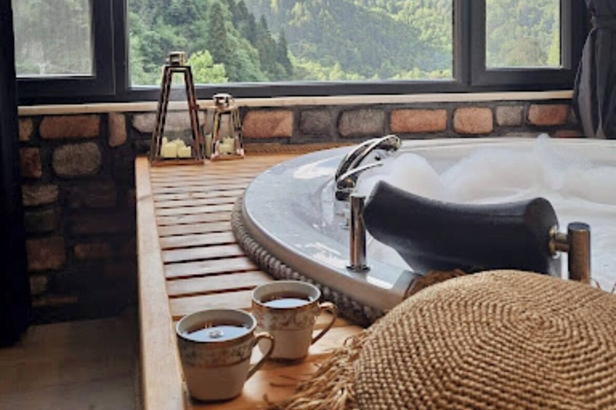 Un cuarto acogedor con un ventanal grande que da a una cadena montañosa boscosa. Un banco de madera recorre la ventana, y una bañera es visible en primer plano.