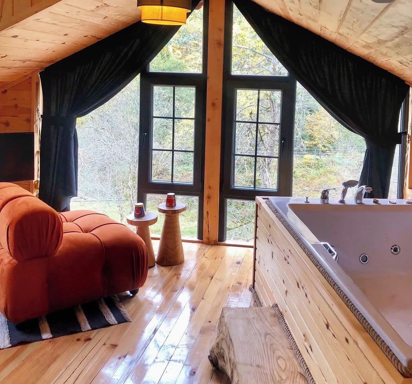 A wooden cabin interior with a large window, a red sofa, a wooden table, and a bathtub.