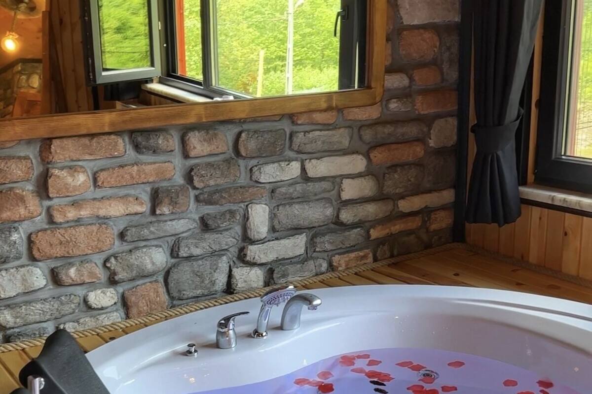 Una habitación de paredes de piedra con una bañera de madera llena de pétalos de rosa, una ventana con vistas a la vegetación y una mesa de madera con dos botellas.