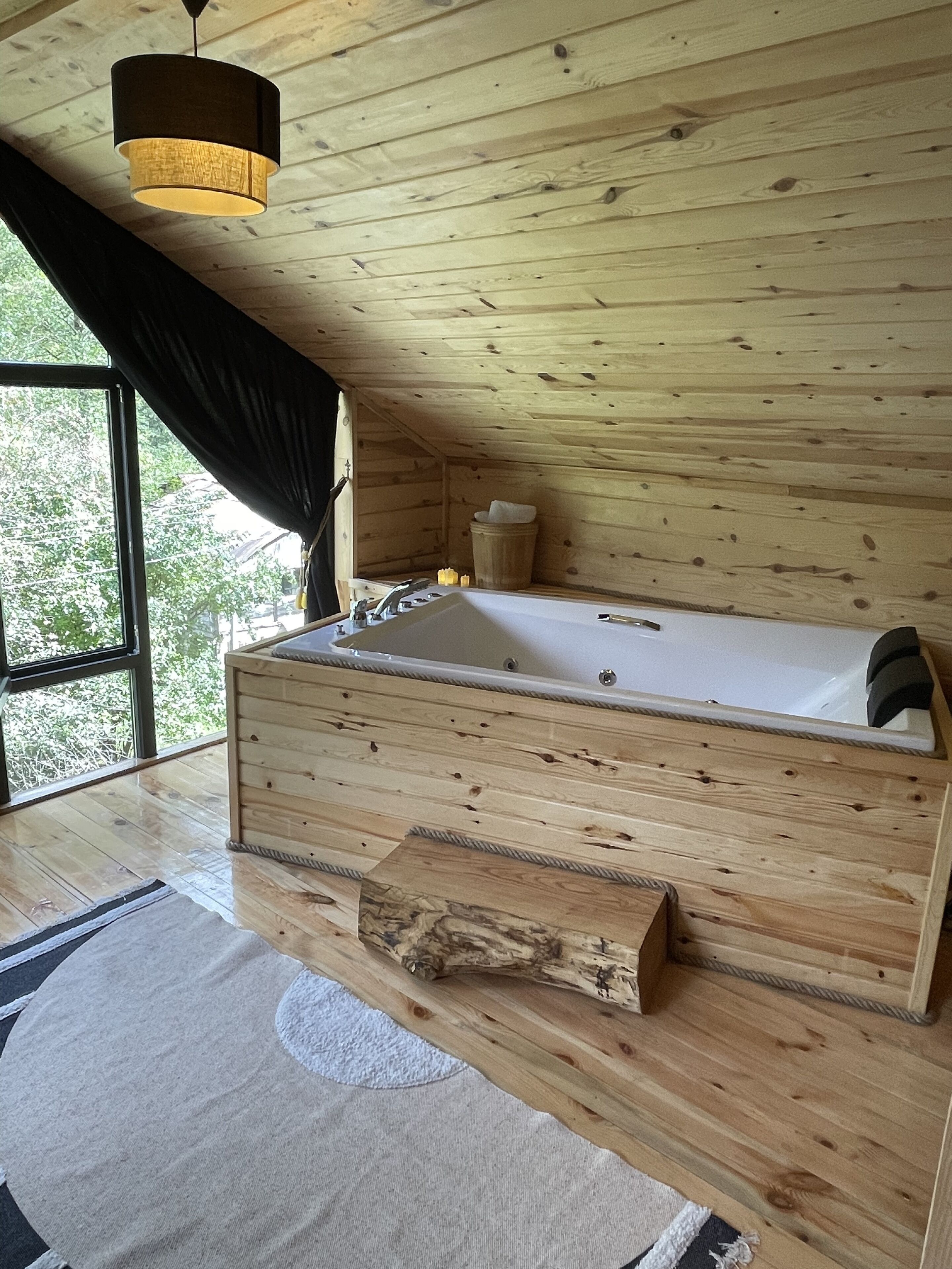 A wooden cabin interior with a large wooden bathtub, a window with a view of trees, and a hanging light fixture.