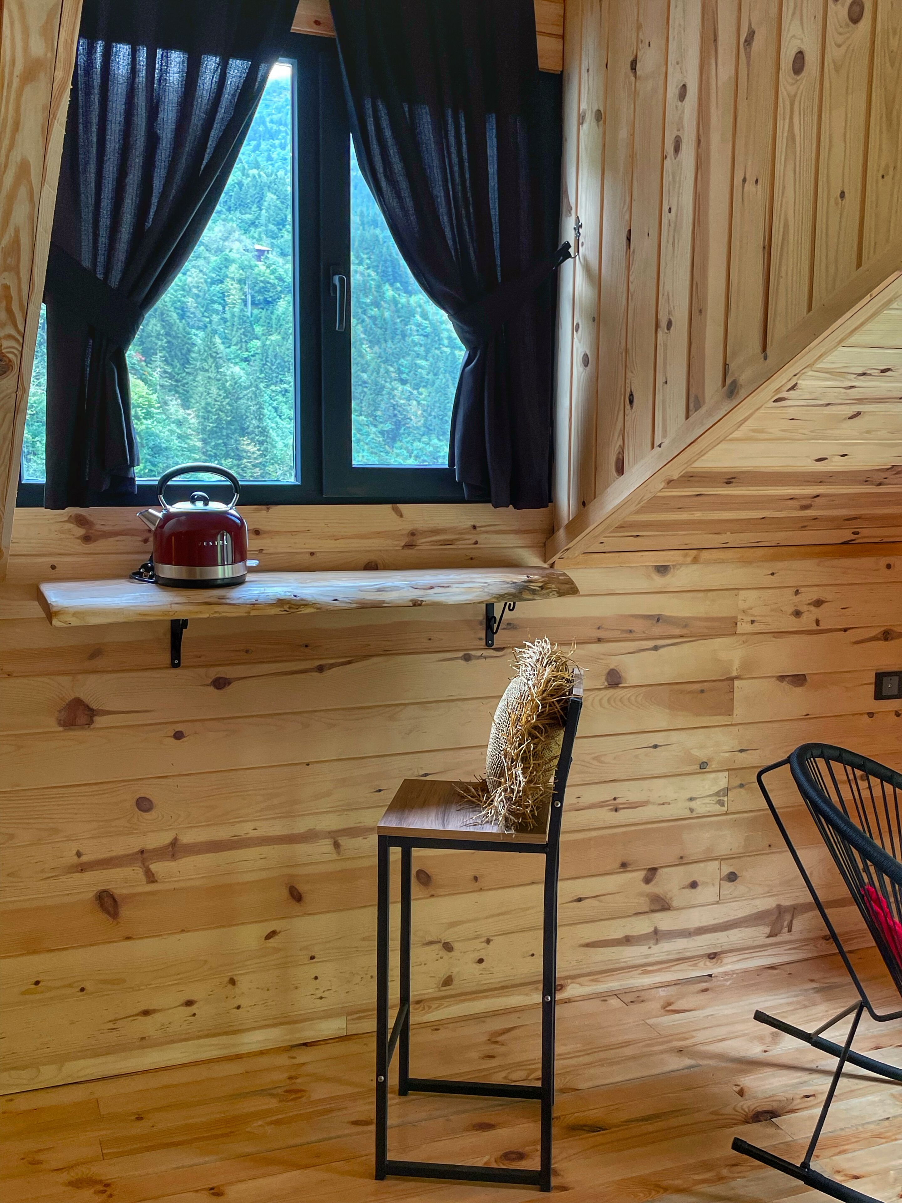 Interior de una cabaña de madera con una ventana, una tetera roja en un estante, una silla con cojín de paja y una silla de metal negro.