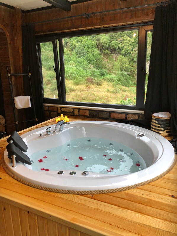 Una cabaña de madera con un ventanal grande, una terraza de madera y un jacuzzi con pétalos de rosa.