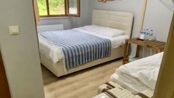 A bedroom with a bed, a window, a wooden headboard, a bedside table with water bottles, and a suitcase.