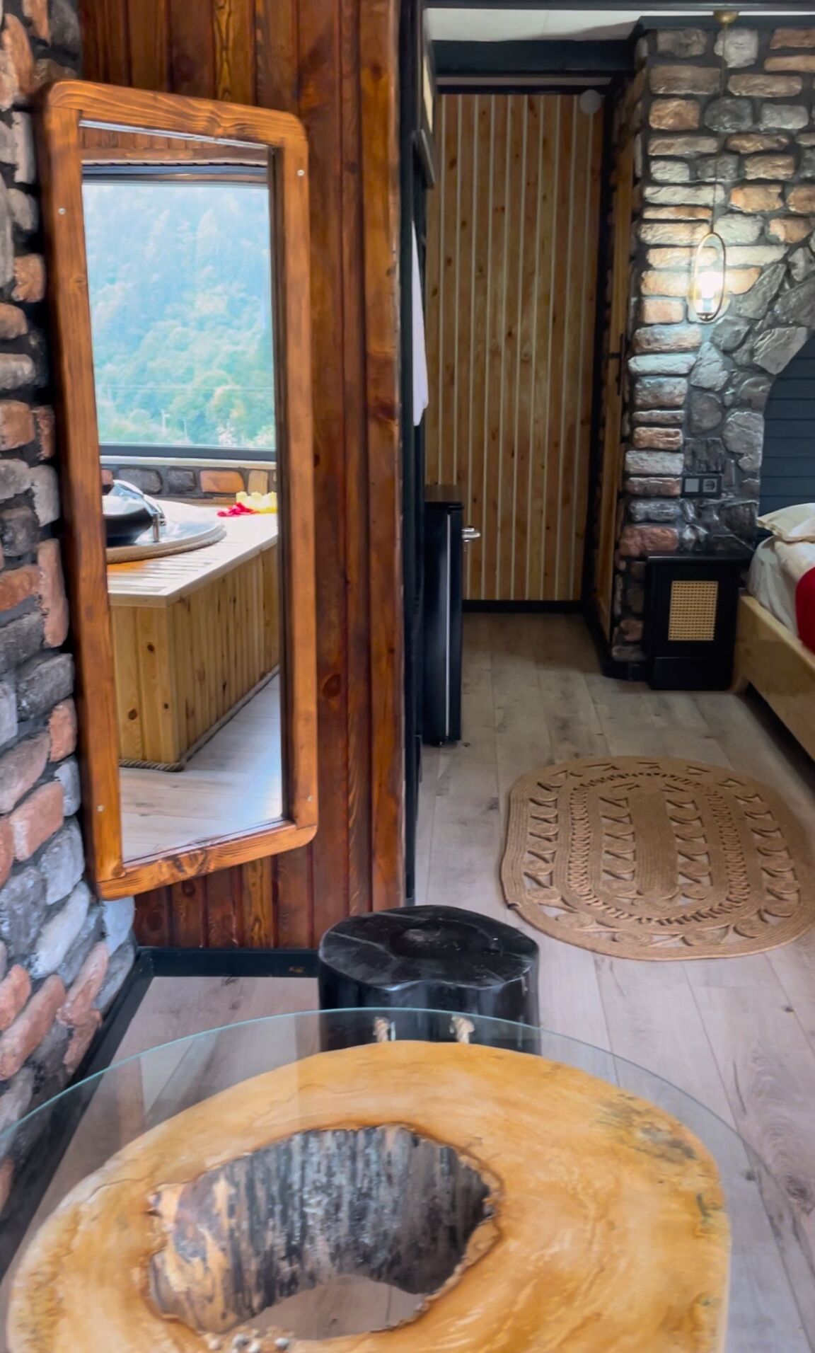 Een rustieke kamer met een stenen muur, houten lambrisering, een grote spiegel, een houten bankje, een zwarte prullenbak en een rond vloerkleed.