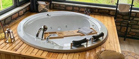 A wooden bathtub with a stone wall backdrop and a view of greenery outside.