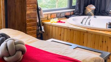 A wooden bed with a red blanket, a wooden bench, a hot tub, and a window with a view of trees.