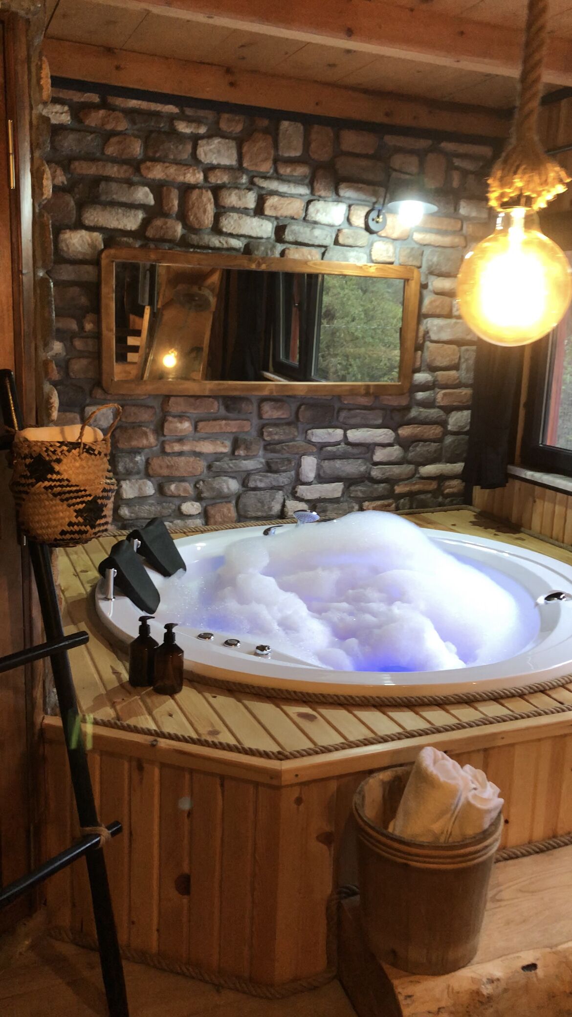 A wooden cabin interior with a stone wall, a wooden bathtub filled with bubbles, a hanging light fixture, and a woven basket.