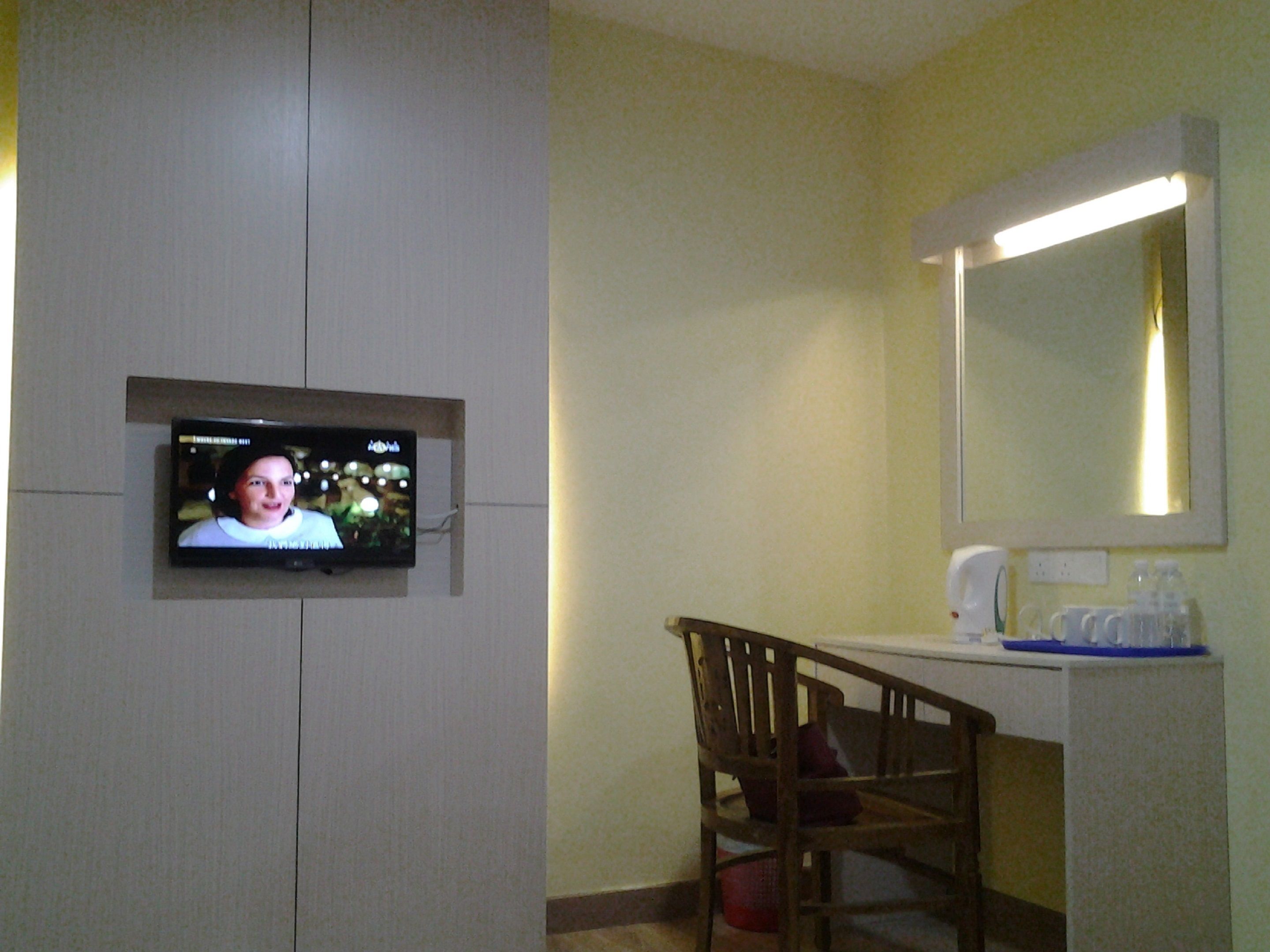Una habitación de hotel con televisión montada en la pared, una silla de madera, un escritorio con espejo y una mesita de noche con artículos de aseo.