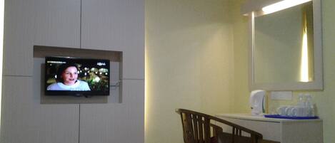 Una habitación de hotel con televisión montada en la pared, una silla de madera, un escritorio con espejo y una mesita de noche con artículos de aseo.
