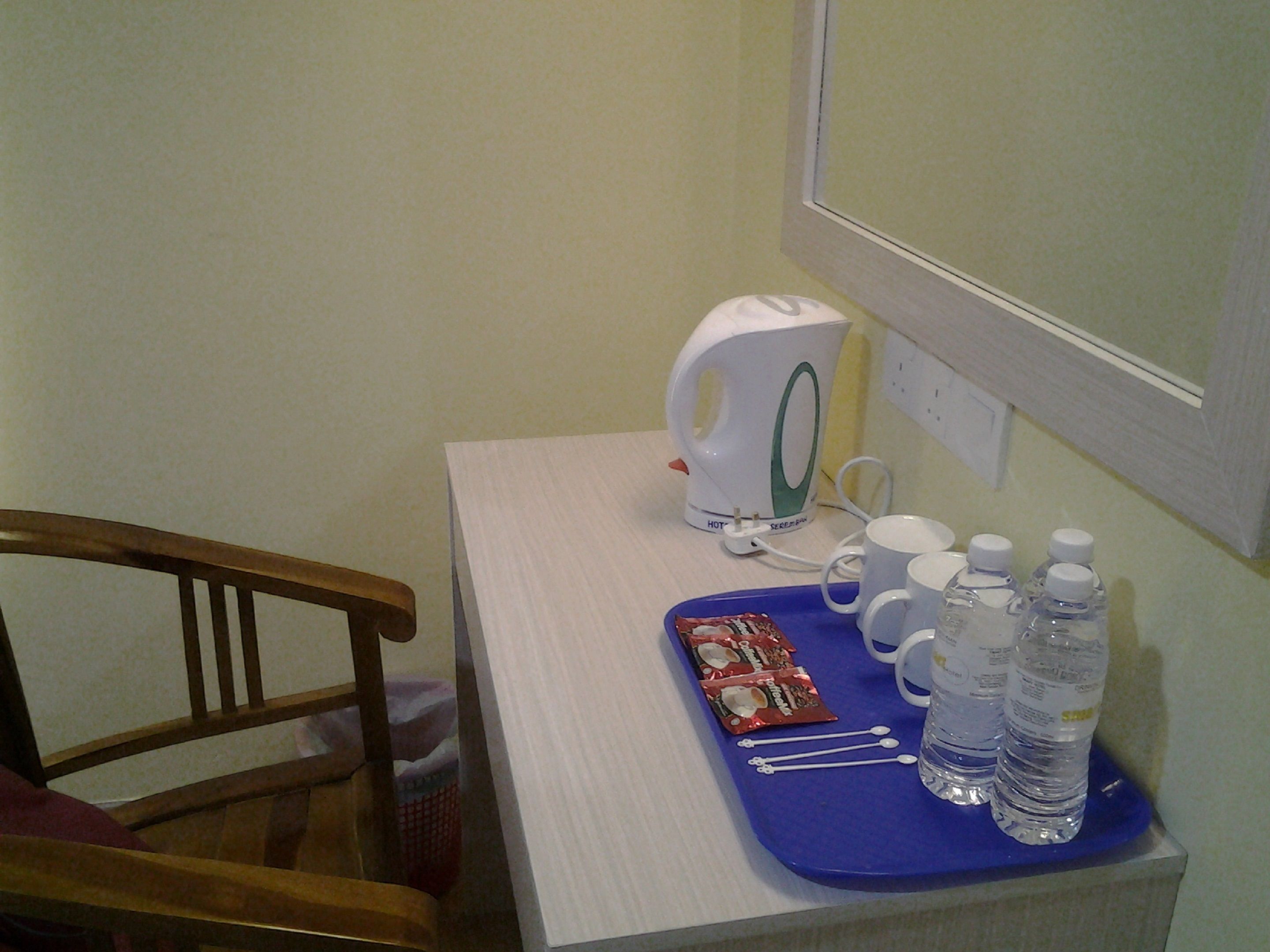 A hotel room with a wooden chair, a white table, a blue tray with water bottles, a coffee maker, and a packet of coffee.