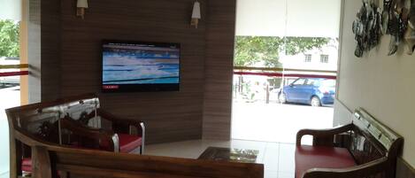 Un hall de hotel con una televisión de pantalla plana grande montada en la pared, muebles de madera y vistas al exterior.