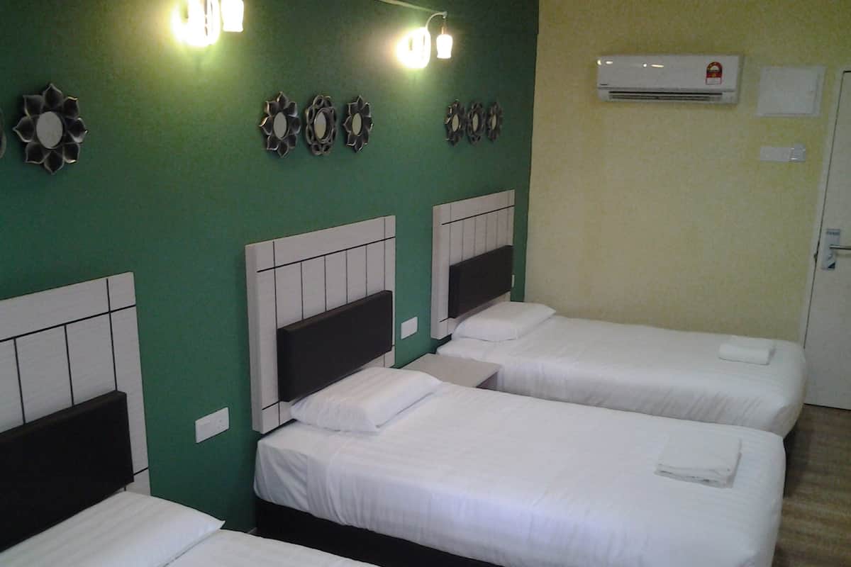 A hotel room with three single beds, each with a white mattress and headboard, against a green wall. There are wall-mounted lights and an air conditioning unit above the beds.