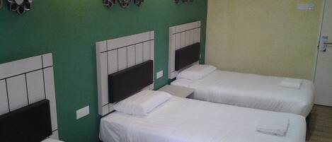 Une chambre d’hôtel avec trois lits simples, chacun doté d’un matelas et d’une tête de lit blancs, le tout adossé à un mur vert. Des lampes murales et un appareil de climatisation sont fixés au mur au-dessus des lits.